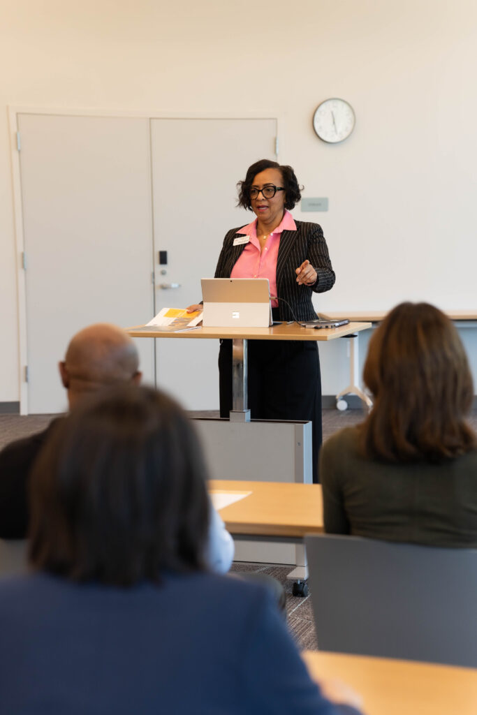 A middle-aged Black woman gestures thoughtfully while teaching a group of adult learners in a bright classroom.