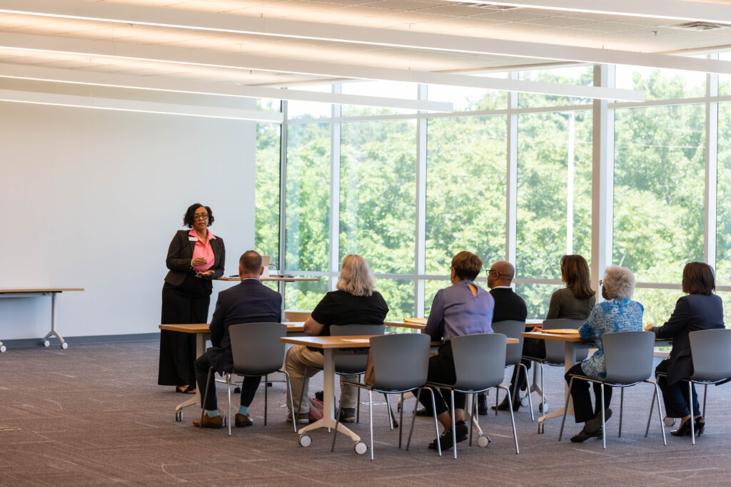 A Black woman teacher stands at the front of a full classroom, engaging a diverse group of adult students in a literacy lesson.