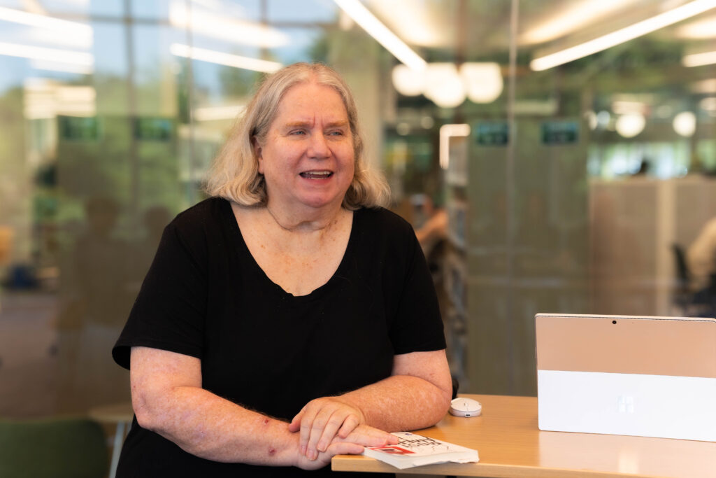 An older white woman stands at the front of a classroom, smiling as she teaches a literacy lesson to a group of adult learners.