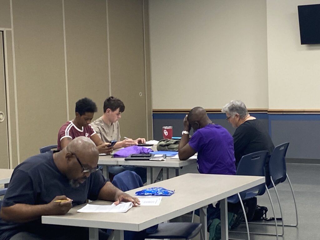 Several adult students are seated at separate tables, focused on their literacy studies in a bright classroom.